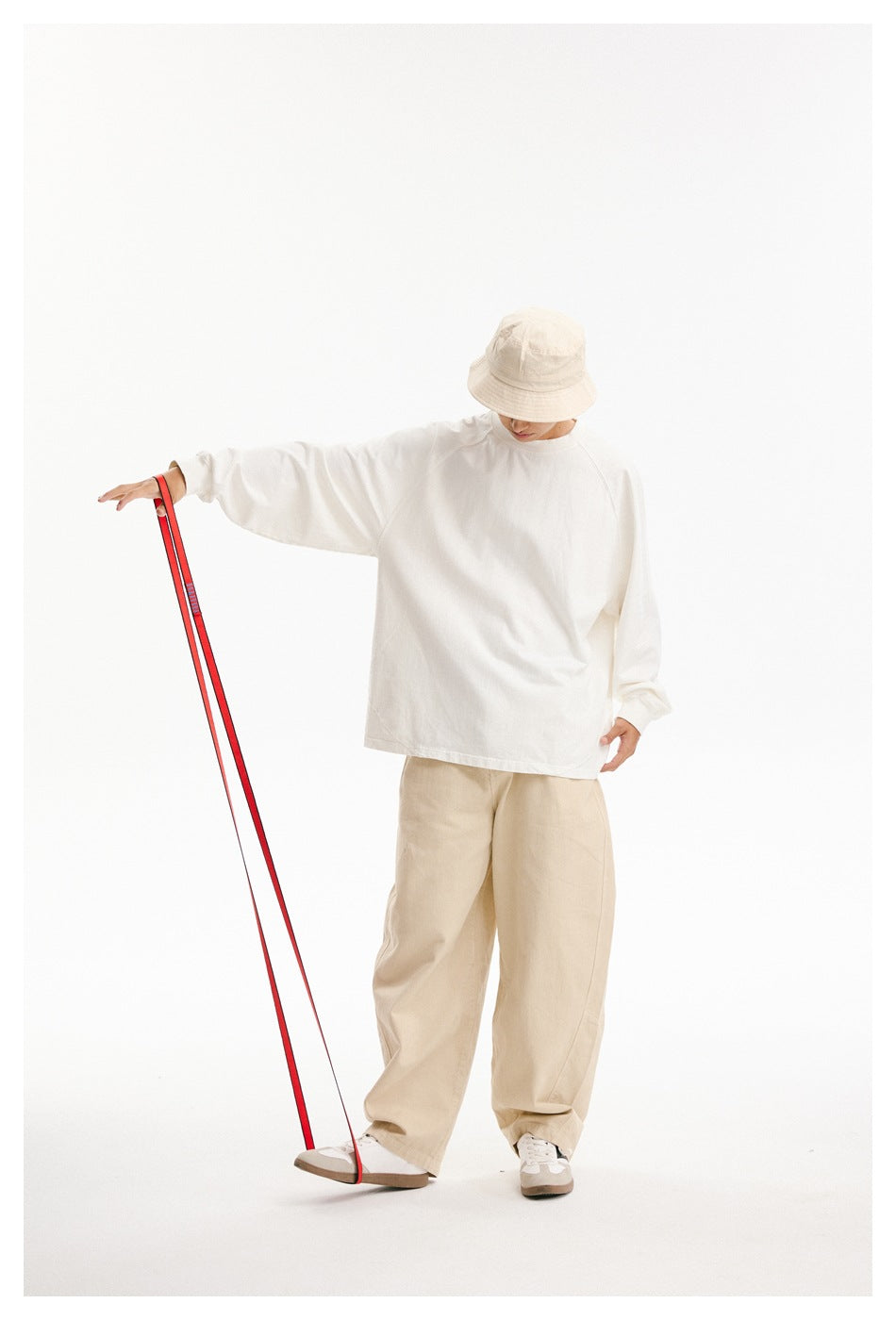 Model in a loose white long-sleeve shirt and beige trousers, holding a red strap beside a beige bucket hat.