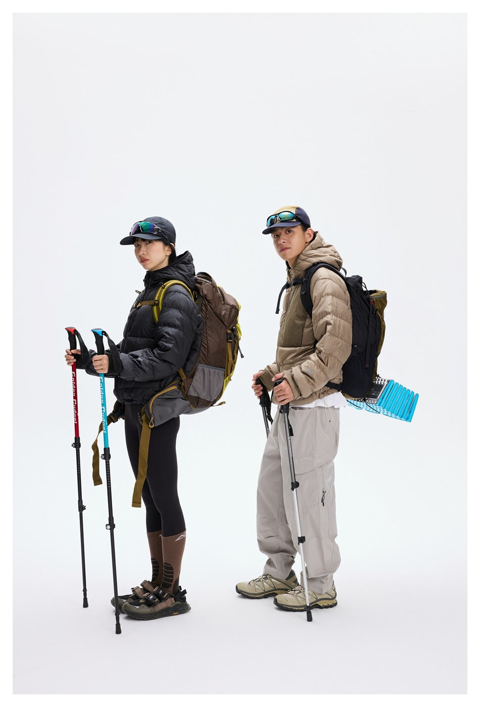 Two hikers pose with trekking poles: one wears a black puffer jacket and the other a taupe jacket, both with backpacks.