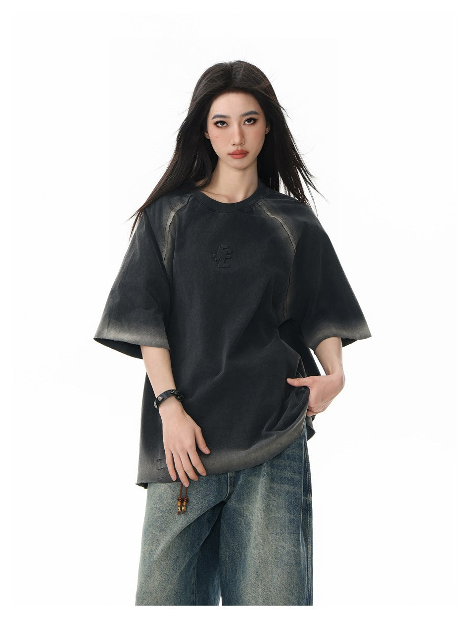 Model wearing an oversized charcoal T-shirt with faded gray accents, raglan seams, and a small raised chest emblem.