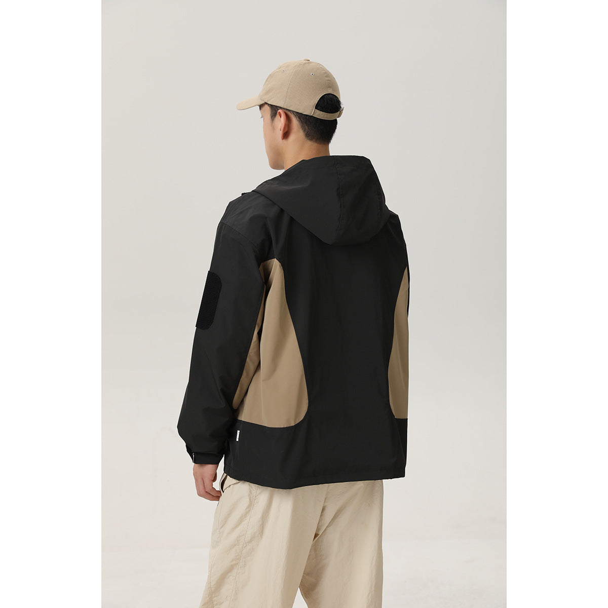 Rear view of a model wearing a black hooded jacket with tan side panels and beige trousers.