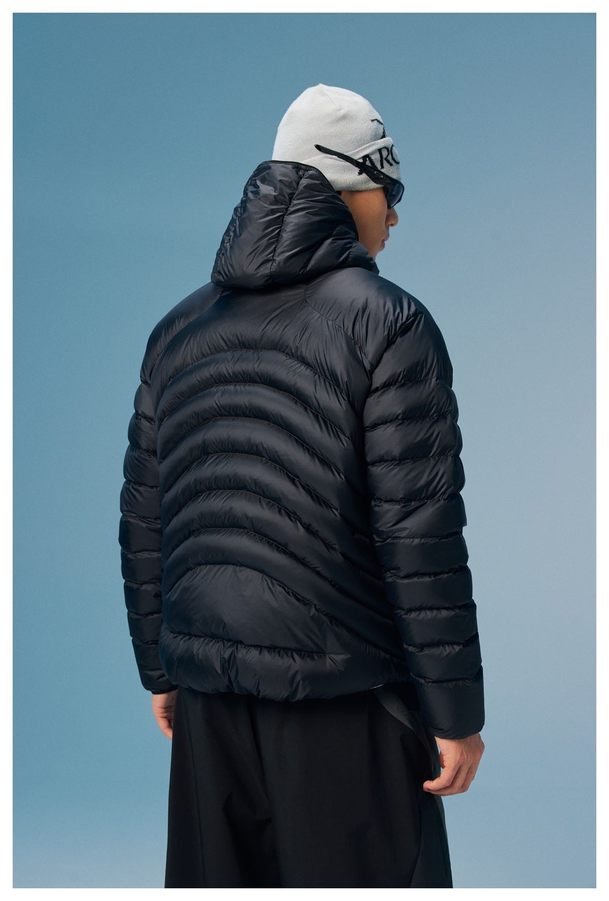 Rear view of a model wearing a black quilted puffer jacket with a large raised hood and curved back quilting.