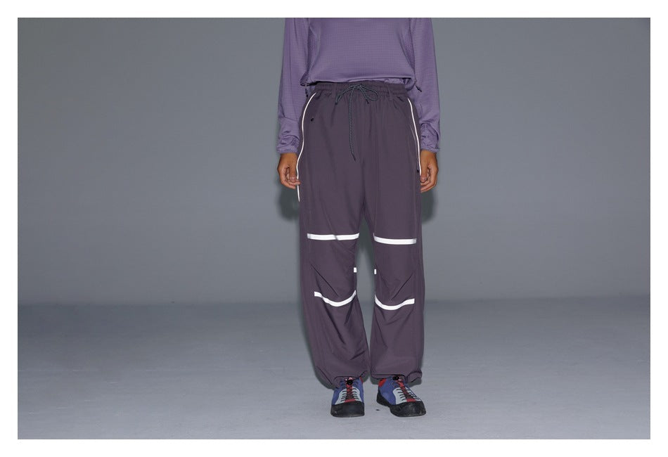Front view of a model wearing dark plum loose pants with white reflective bands, side piping and a drawstring waist.