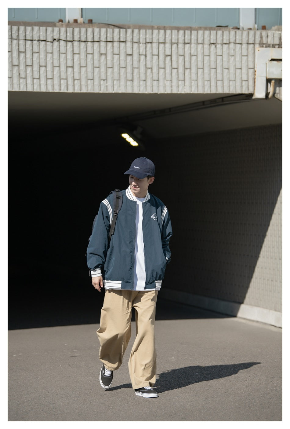 A model walks in a dark teal jacket with striped shoulders and trim, worn over a white shirt.
