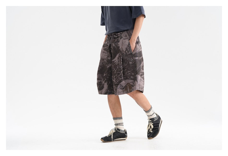 Model wearing loose charcoal-gray graphic shorts, shown from the front at an angle in a wide, white studio scene.