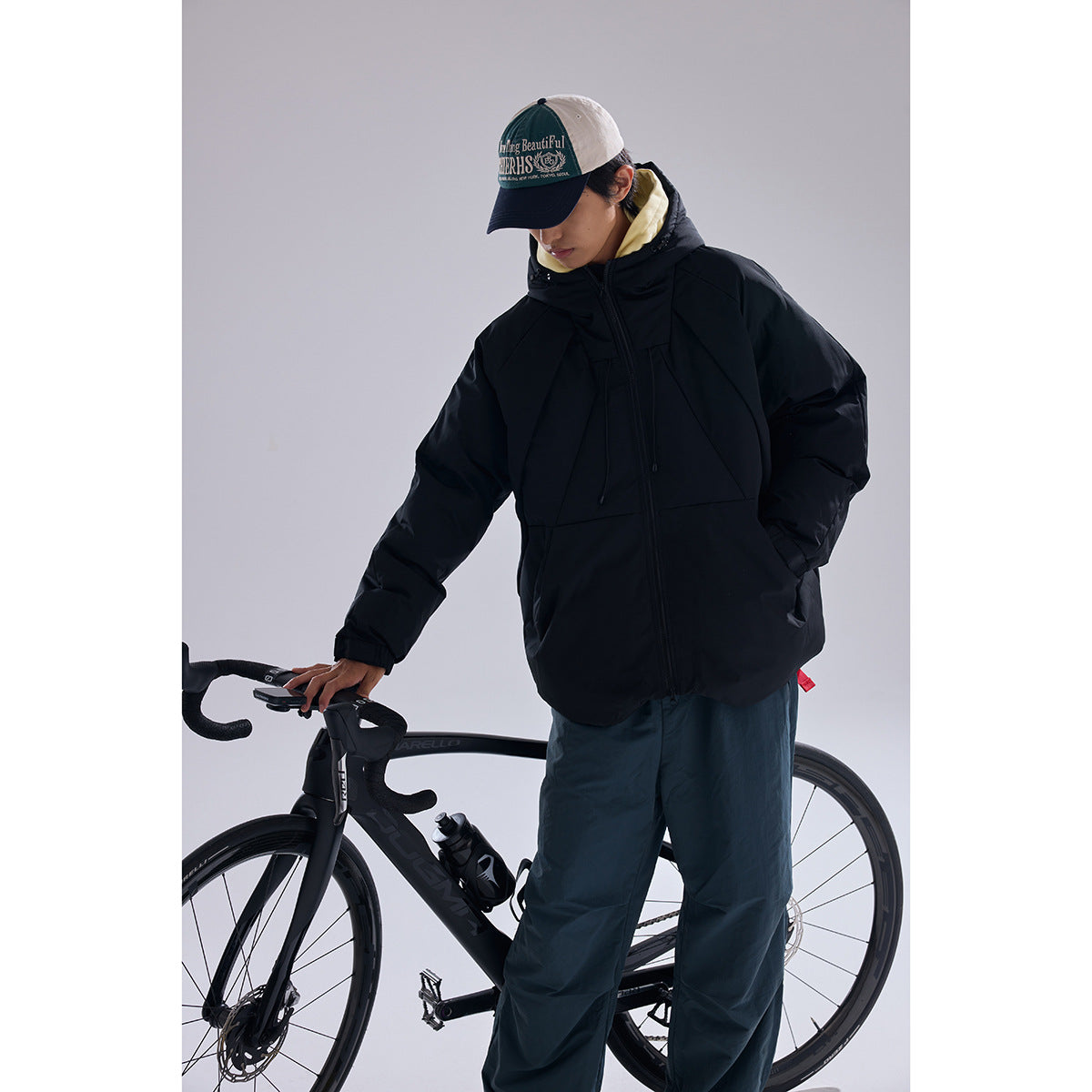 Model wearing a black hooded puffer jacket and dark pants beside a road bicycle, shown from the front at an angle.