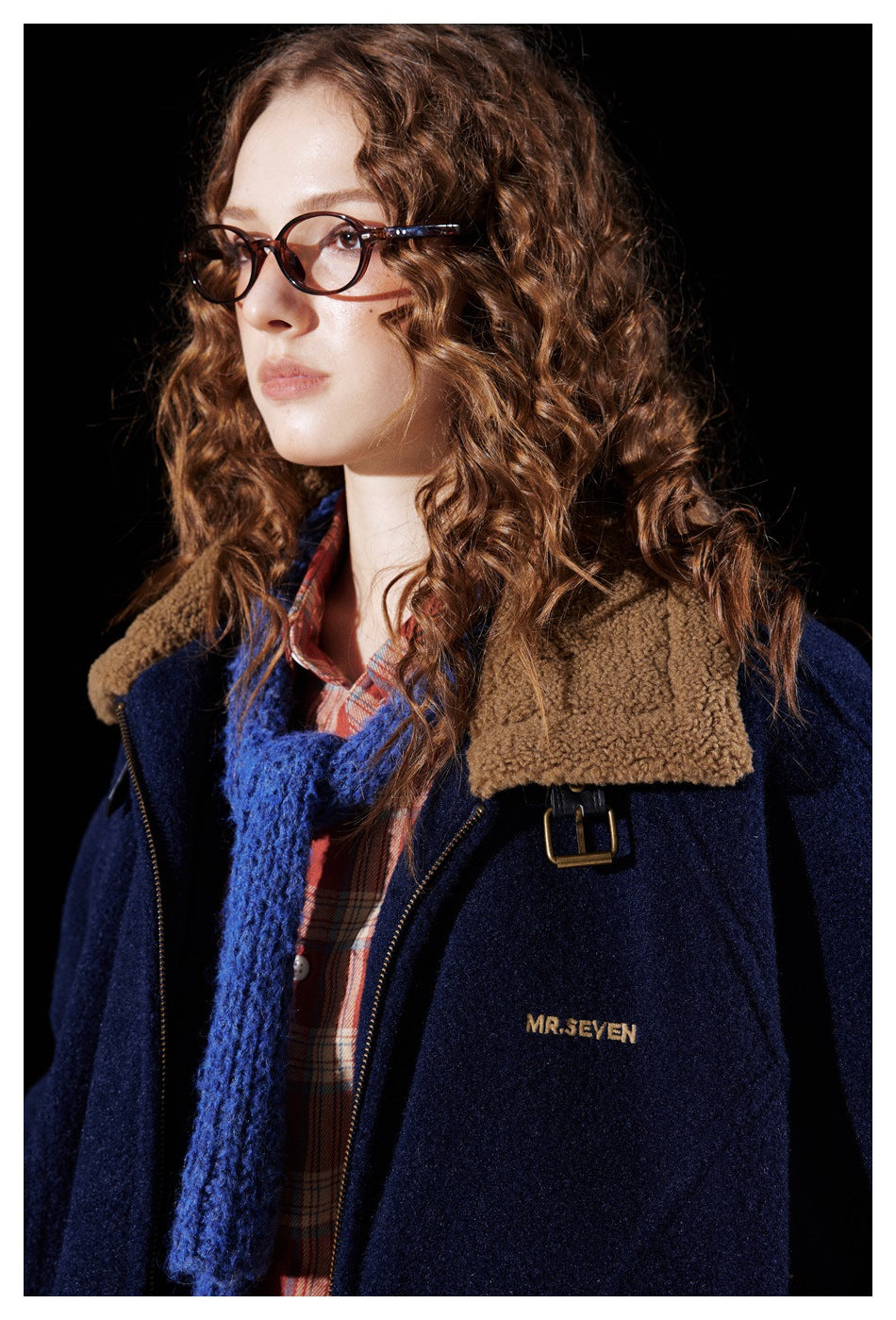 Model in a navy jacket with a tan fleece-like folded collar, buckle strap and zipper, worn over a plaid shirt and blue scarf