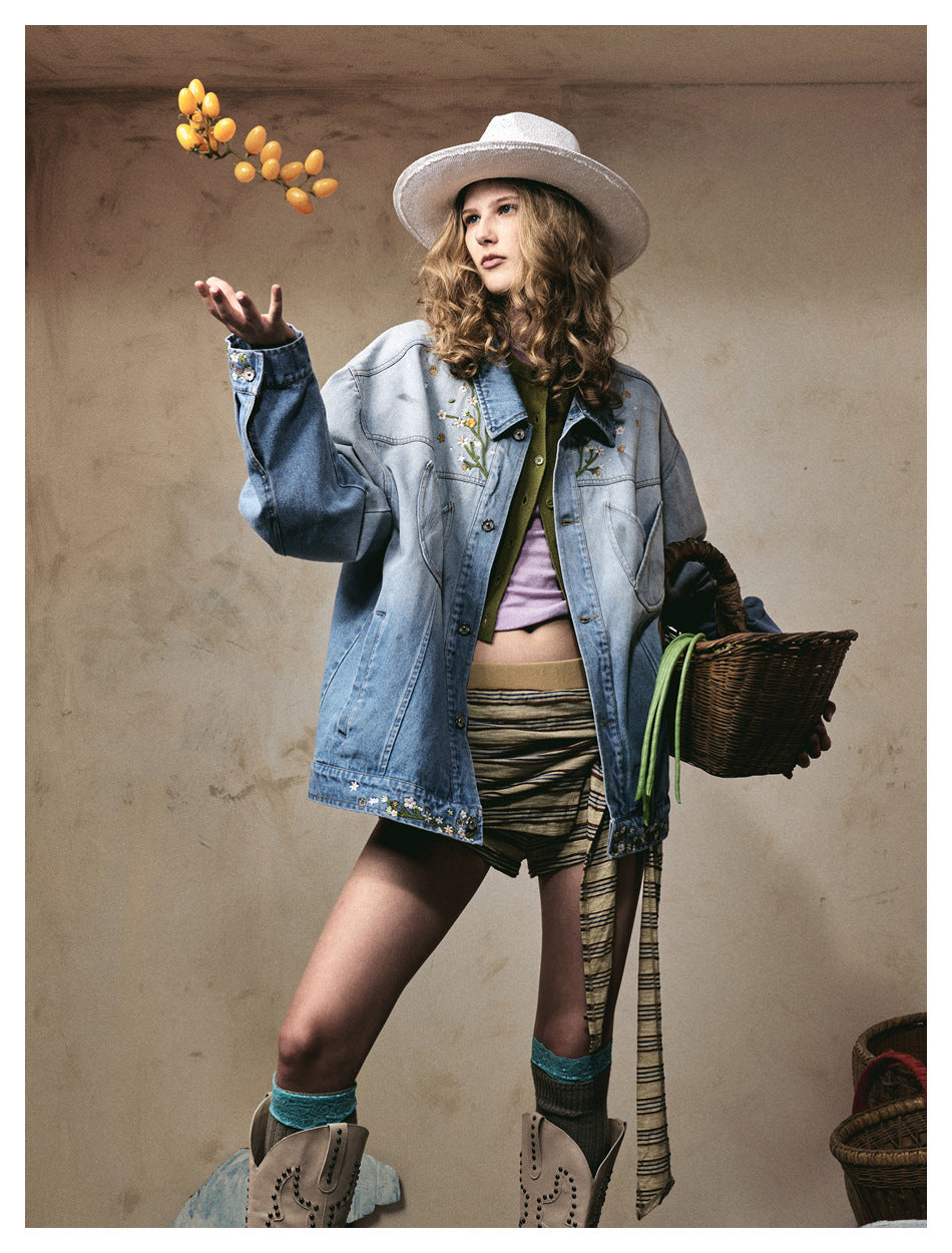 Model wearing an oversized light blue floral-embroidered denim jacket, posing with a basket and wearing a white hat.