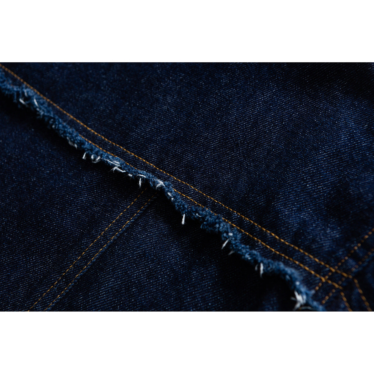 Macro view of dark indigo denim with a diagonal frayed seam and gold stitching.