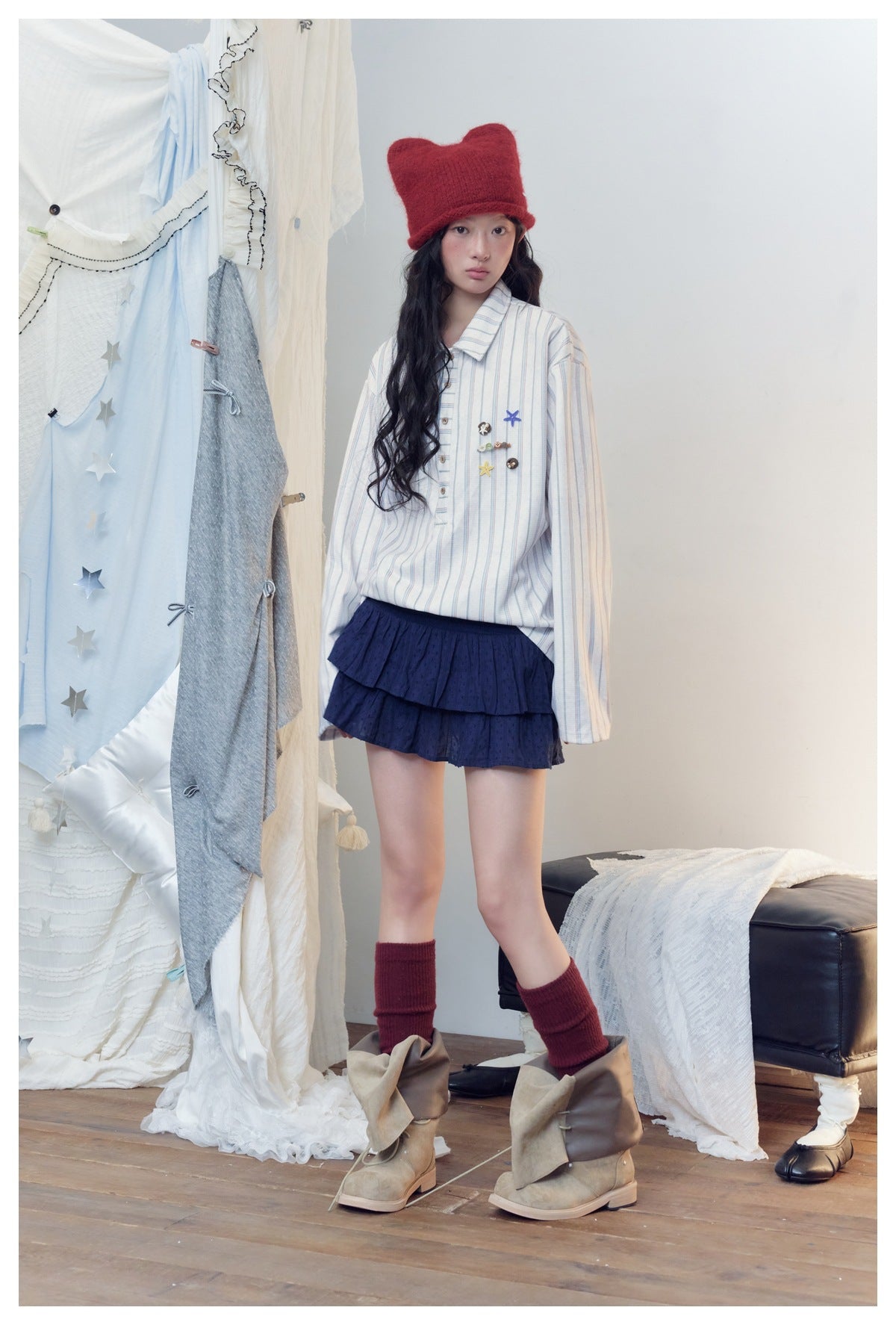 Model wearing a white shirt with fine blue and red stripes and decorative charms, a navy tiered skirt, and a red knit hat.