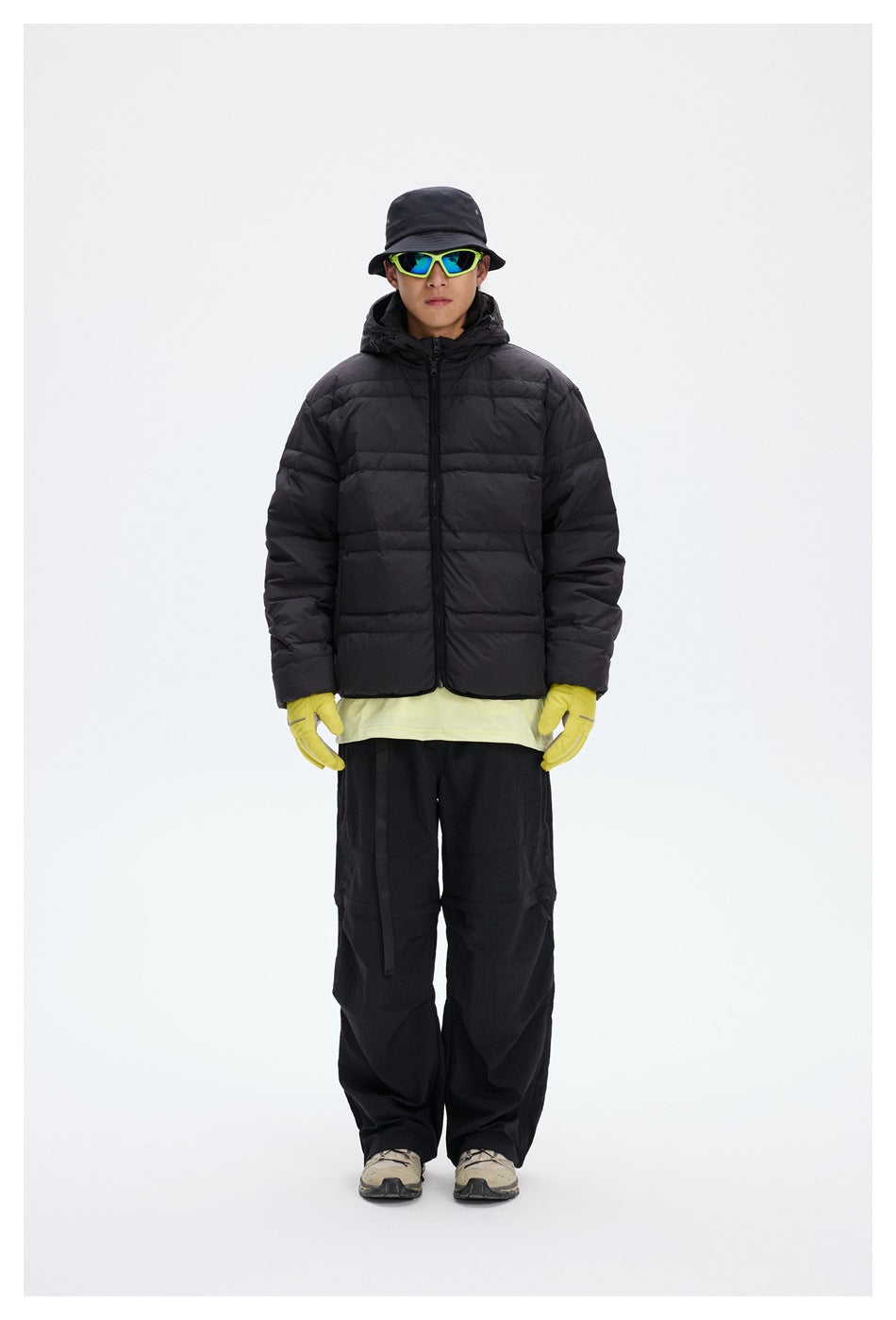 Model in a black quilted hooded jacket with a front zip, black trousers, yellow gloves, bucket hat and sunglasses.