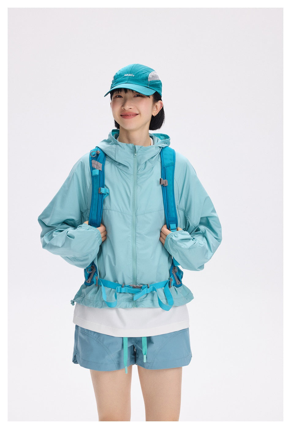 Model wearing a pale aqua hooded jacket, blue hiking backpack, teal cap, and matching shorts, front view.