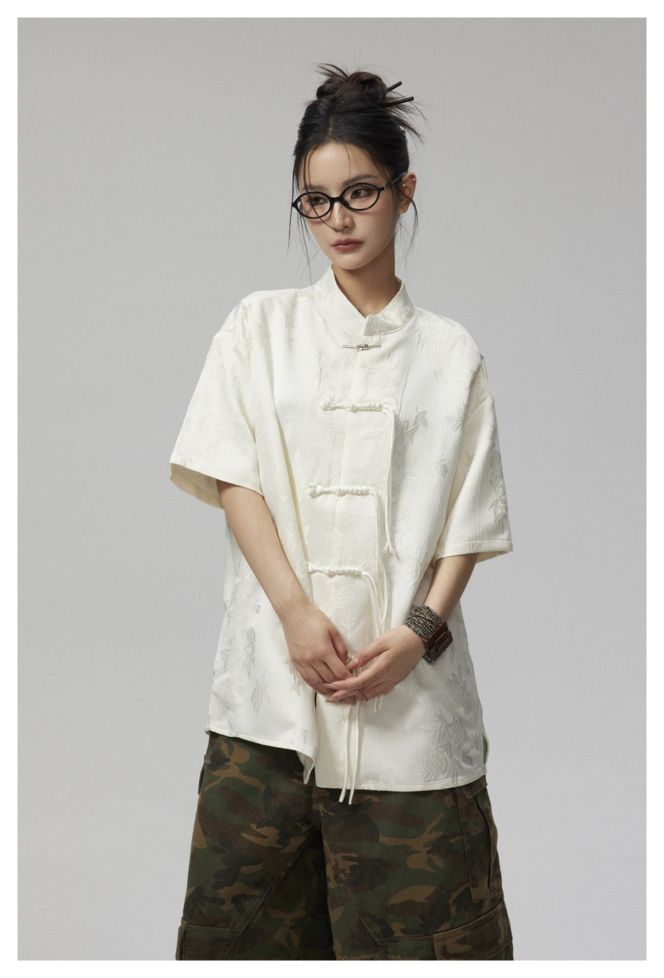 Model wearing an ivory floral short-sleeve shirt with a stand collar and braided closures, paired with camouflage trousers.