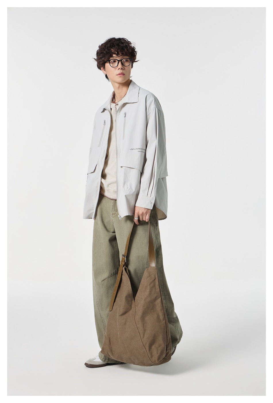 Angled view of a model in a pale gray zip jacket with multiple pockets and loose olive trousers, carrying a brown tote.