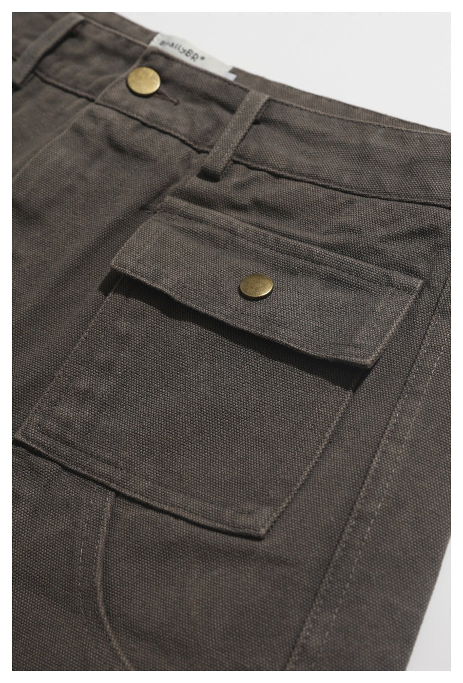 Close-up of charcoal-brown utility trousers showing a flap pocket with a brass-tone snap and waistband button.