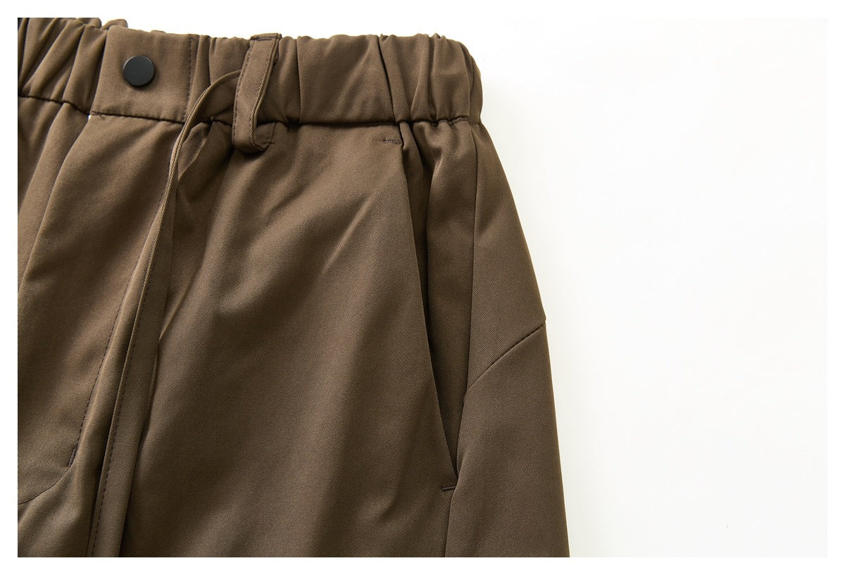 Close-up of brown trousers showing a gathered waistband, black button, belt loop and side pocket.