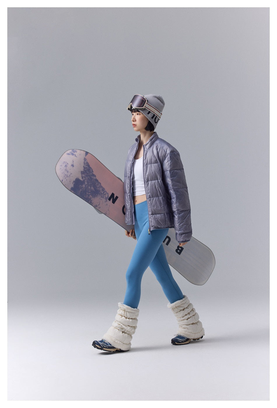 Model walking with a snowboard in a metallic gray quilted jacket and blue leggings.