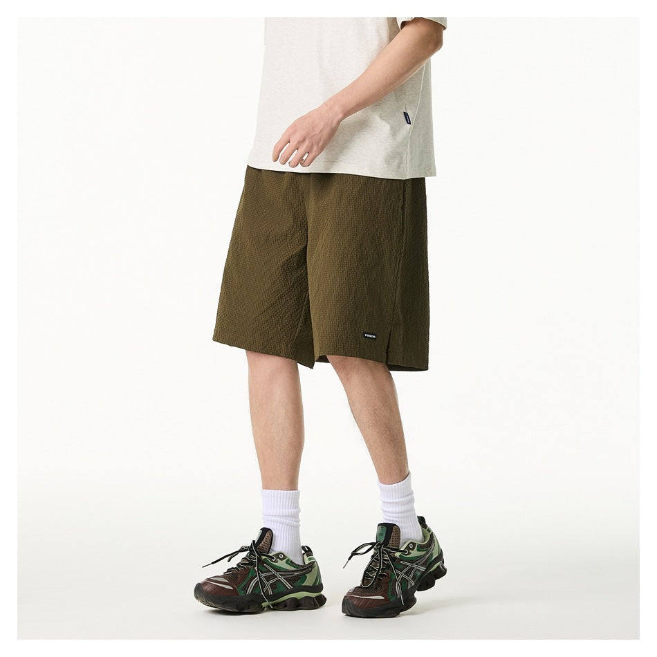Angled side view of a model wearing olive textured knee-length shorts with a light T-shirt and sneakers.