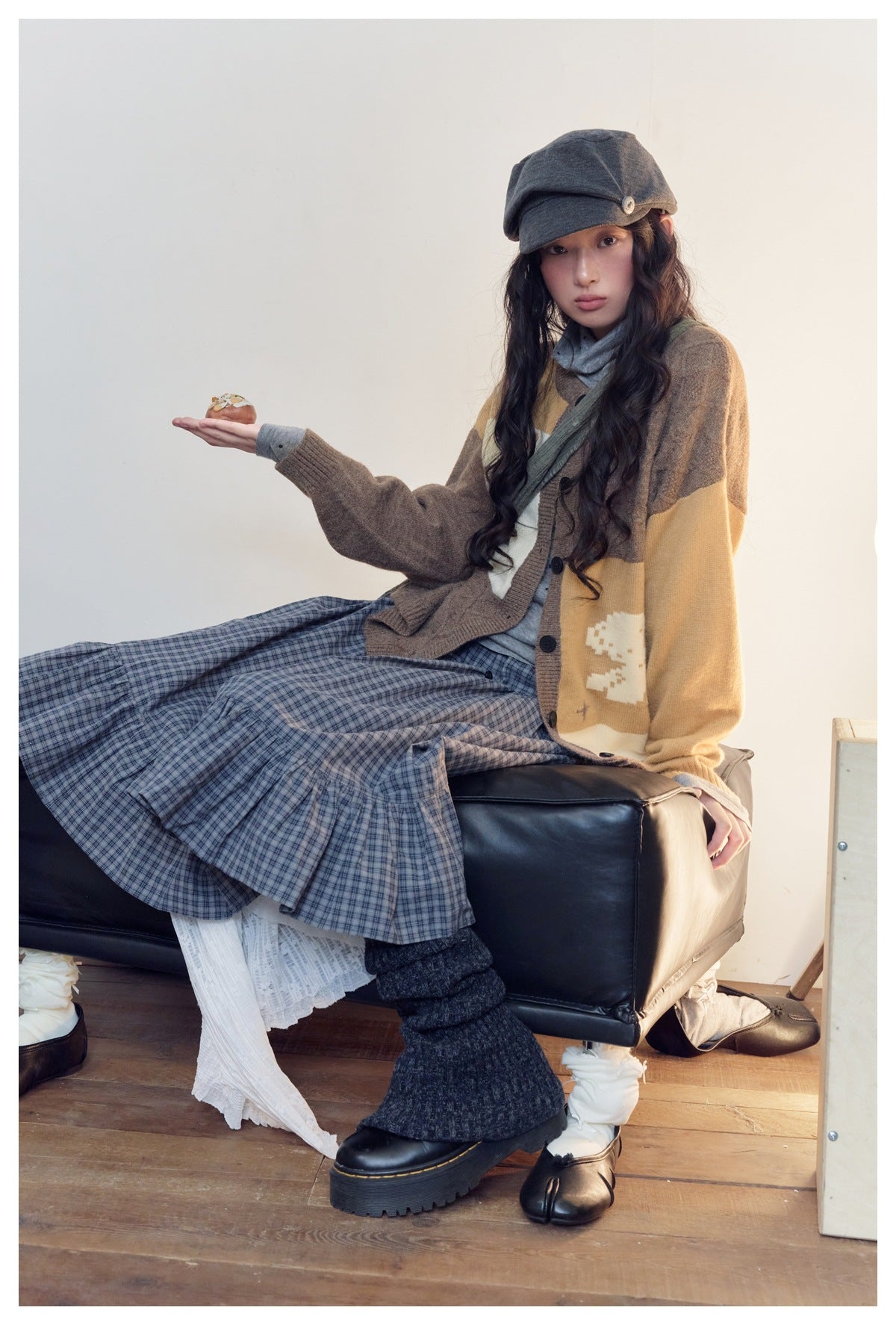 Seated model wearing a brown and mustard graphic cardigan with a blue plaid skirt and gray cap.