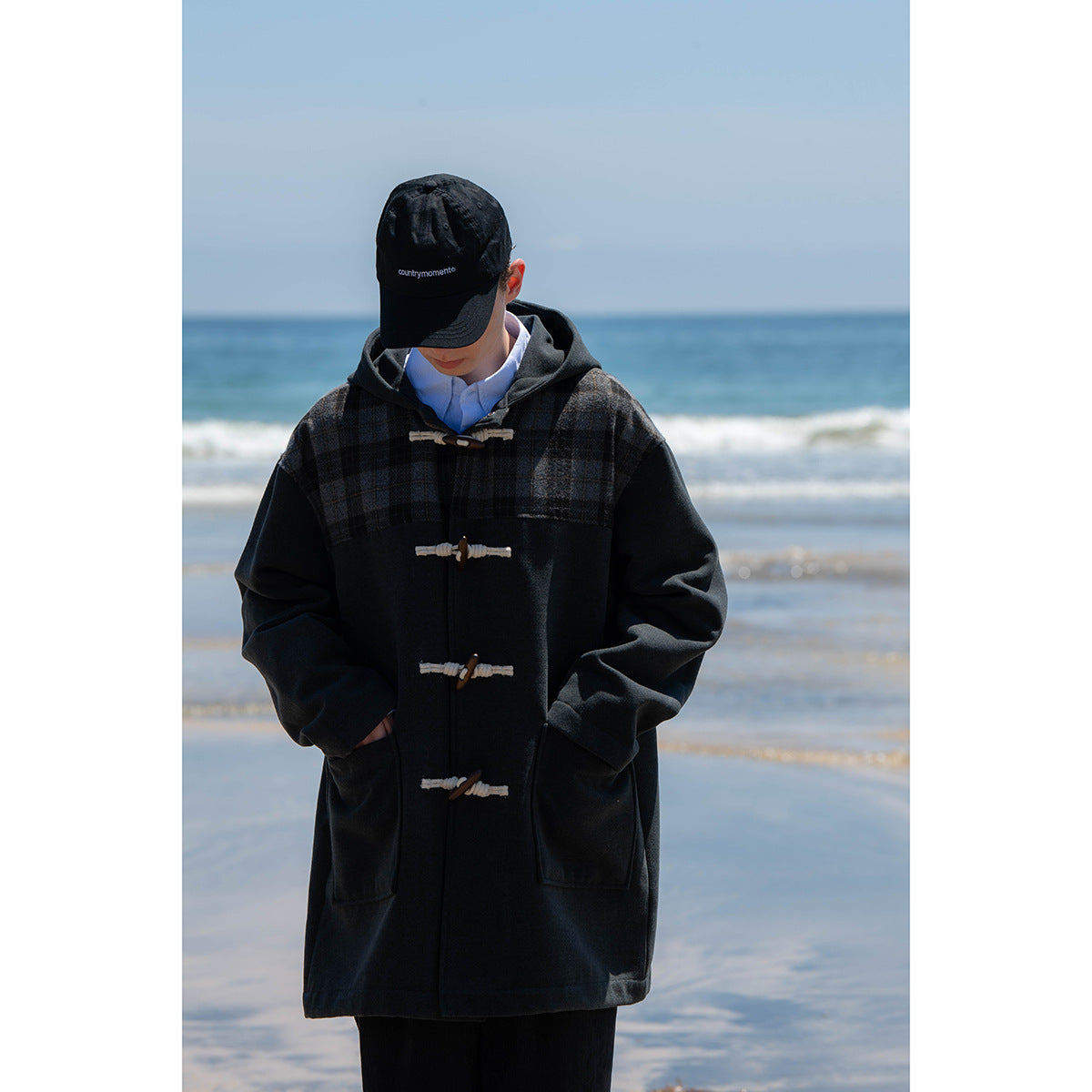 Person wearing a dark hooded duffle coat with charcoal plaid shoulder panels and toggle closures on a beach.