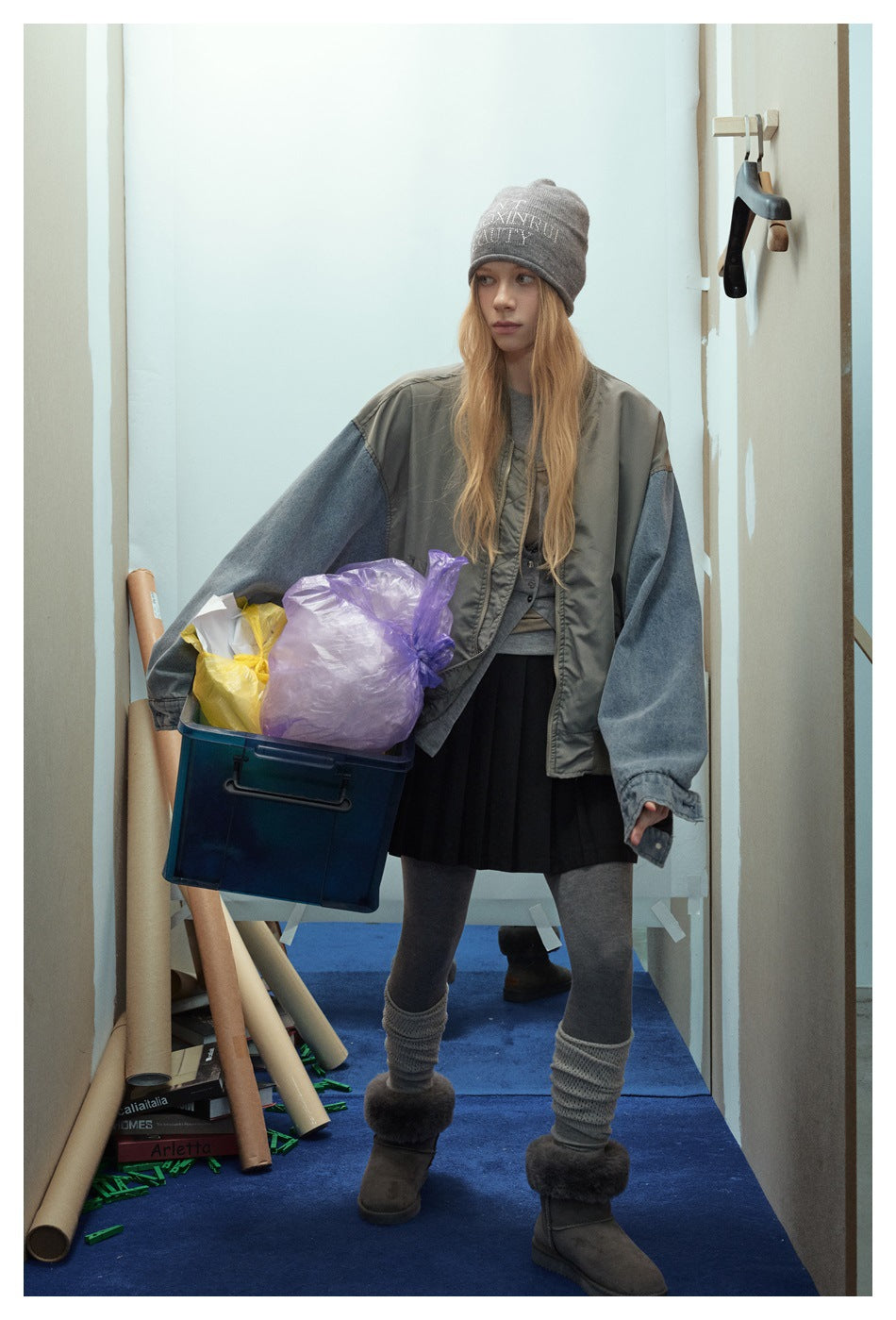 Model wearing an oversized olive-gray jacket with blue denim sleeves, a gray beanie, black pleated skirt and gray leggings.