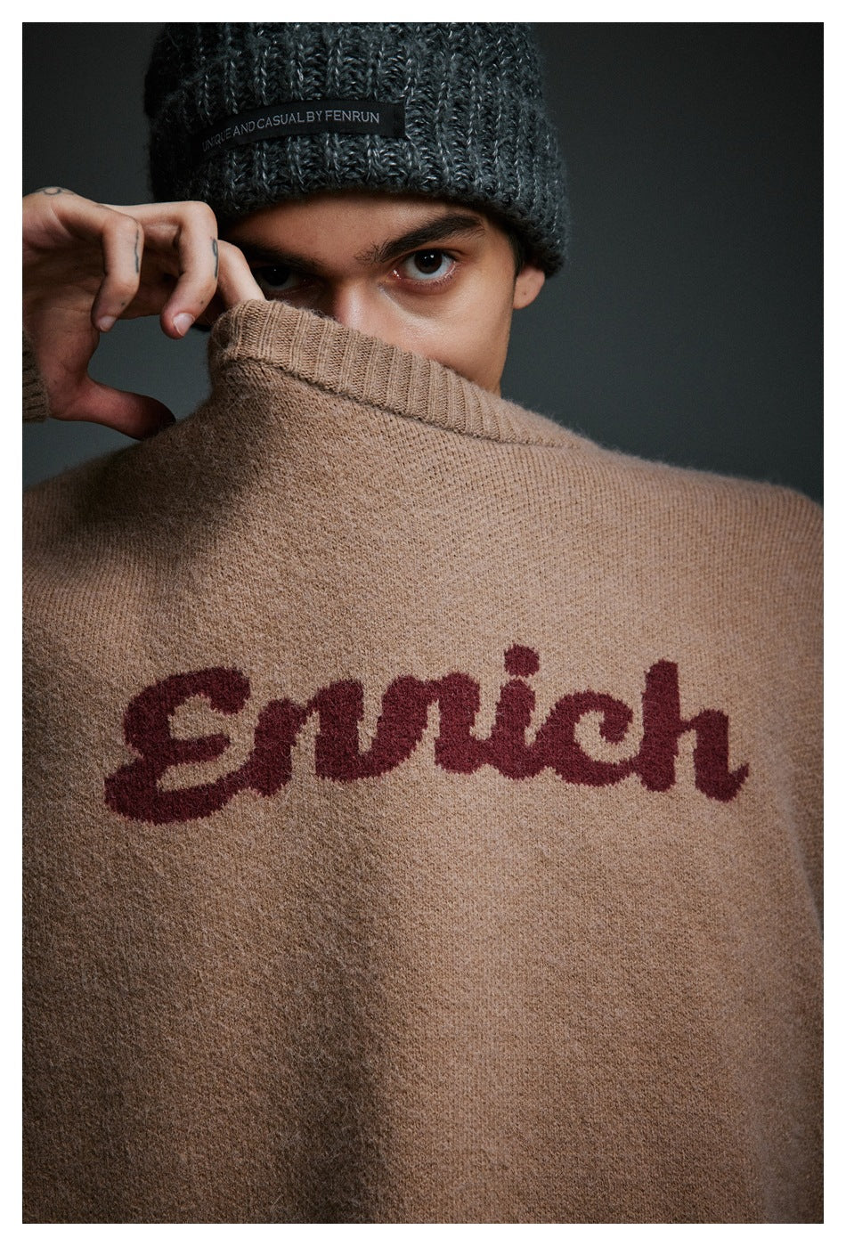 Close-up of a model holding up a taupe sweater to show its burgundy script lettering and ribbed collar.