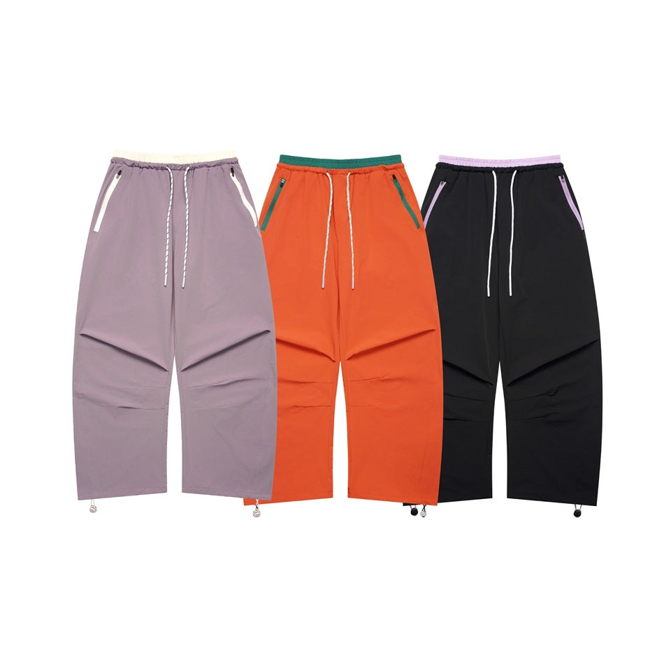 Three pairs of loose pants shown from the front in mauve, orange, and black, with drawstrings and zip pockets.