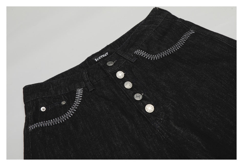 Angled close-up of black jeans with silver fly buttons and white curved stitching around the front pockets.