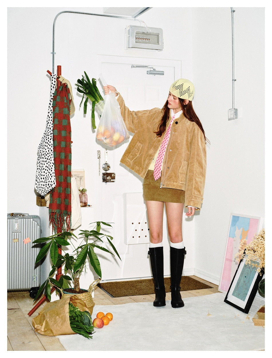 Full-length model in a tan textured jacket, olive mini skirt, pink polka-dot tie, cream knit cap, and black knee-high boots.