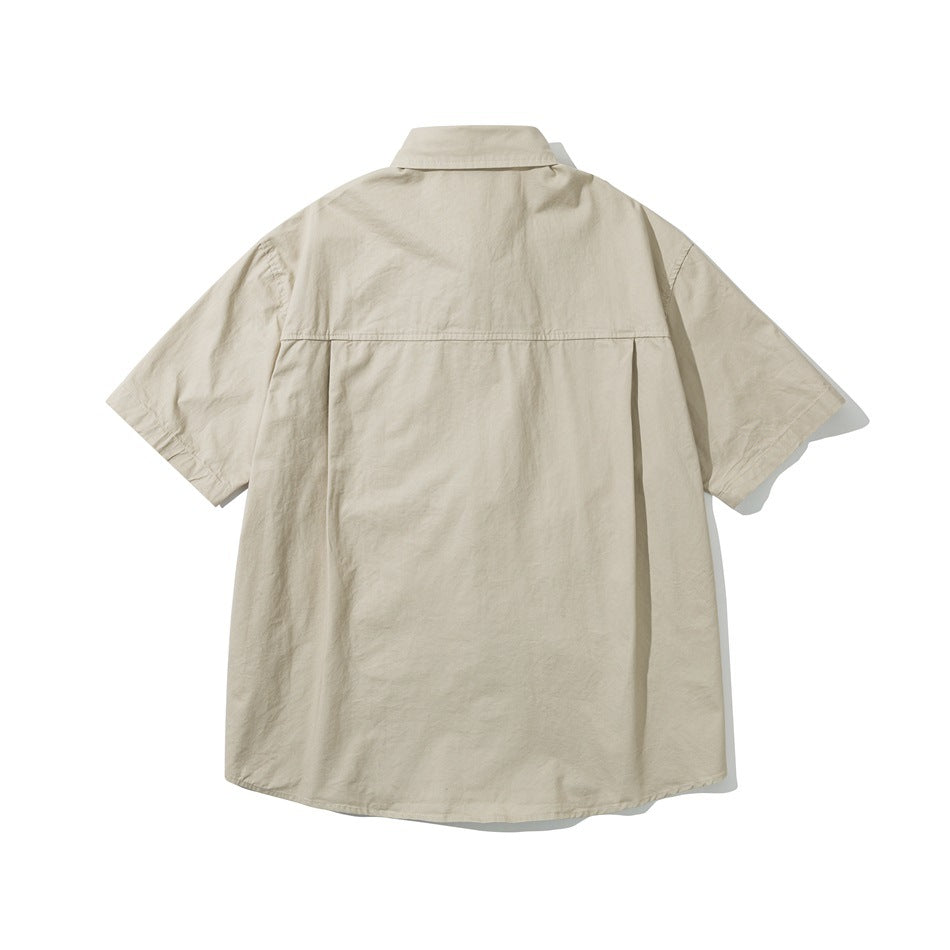 Back of a light khaki short-sleeve shirt showing a yoke seam, center pleat and curved hem, laid flat