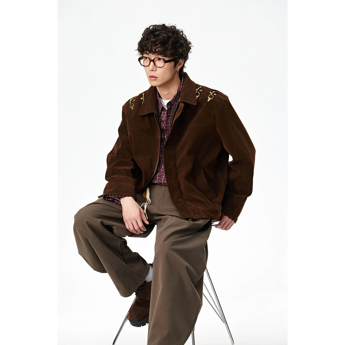 Seated model wearing a dark brown jacket with gold shoulder hardware, plaid shirt, and loose taupe trousers.