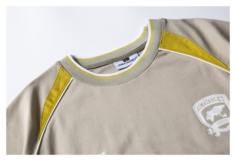 Close-up of a beige crewneck shirt with yellow shoulder inserts, white piping, and a chest crest.