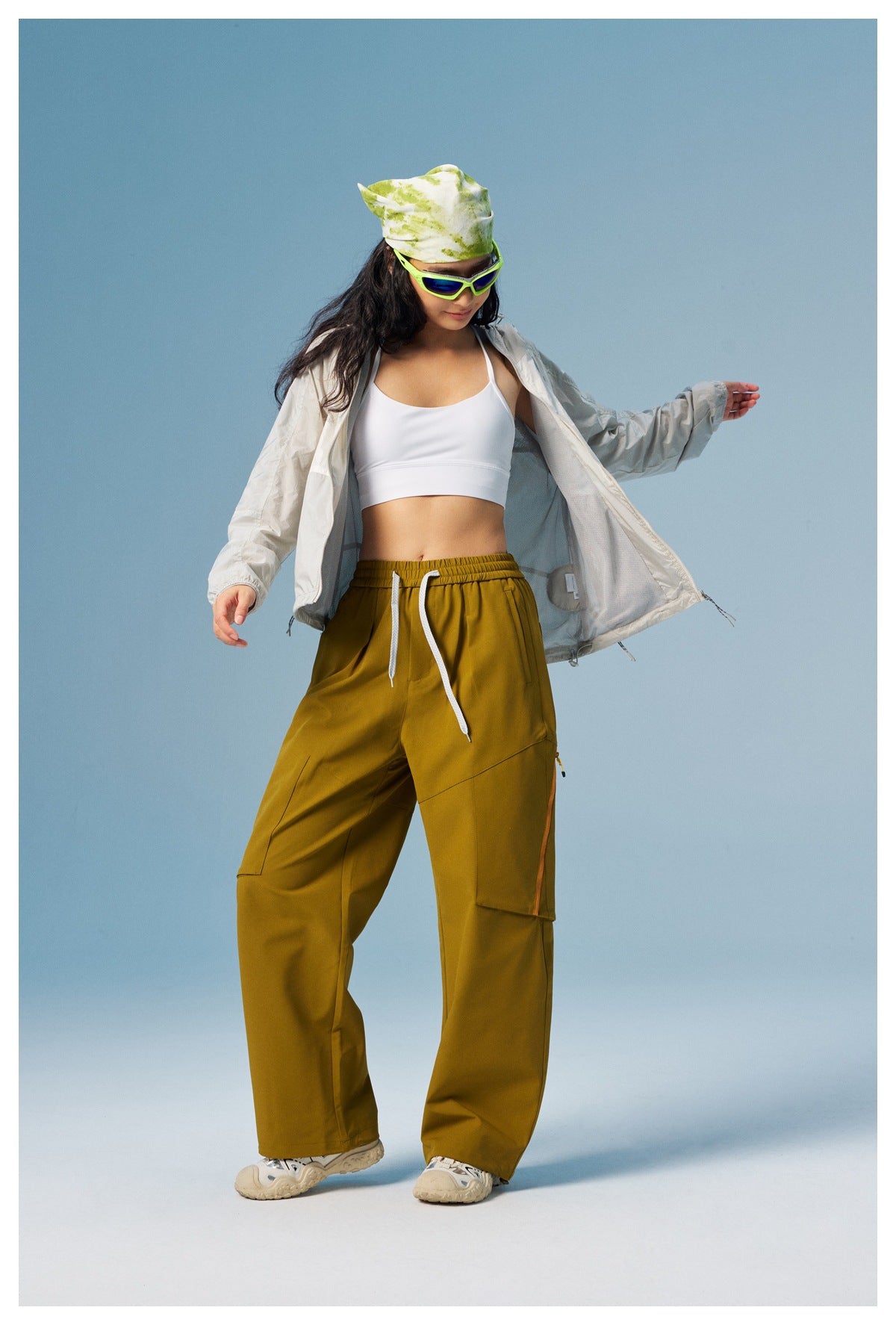 Woman models an open light gray hooded jacket with mustard cargo pants and a white crop top.