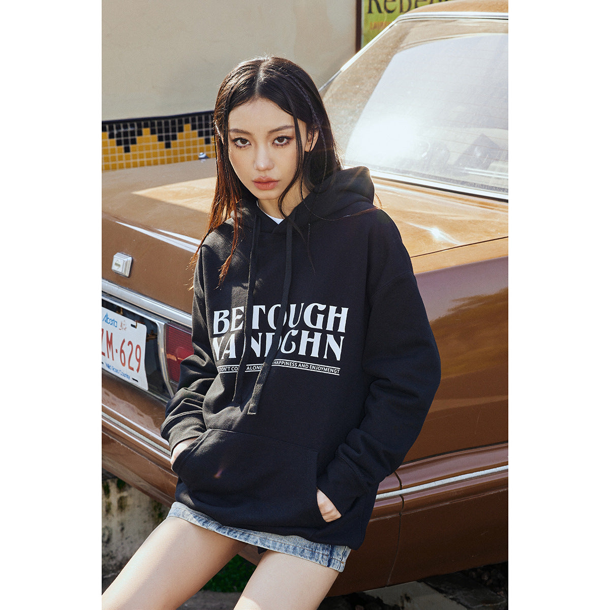 Model wearing an oversized black pullover hoodie with white chest lettering and a kangaroo pocket beside a brown car.