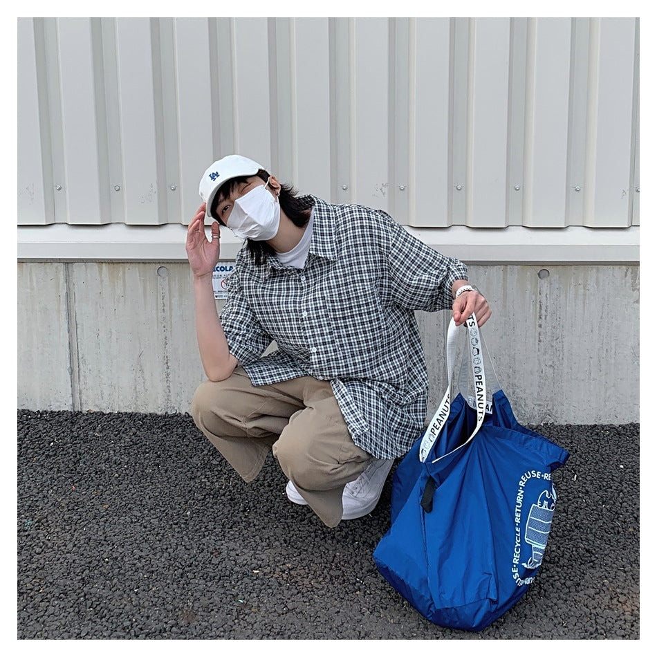 A masked person crouches outdoors wearing a dark plaid short-sleeve button-up shirt and khaki pants.