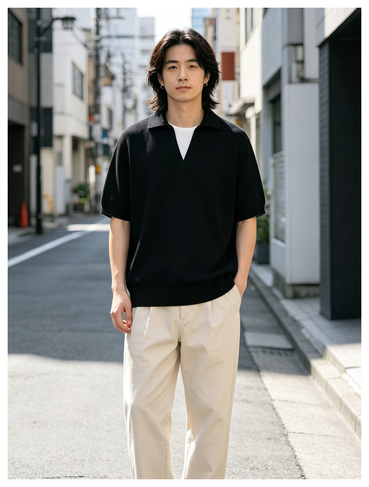 Man standing on a city street wearing a black short-sleeve knit polo with open V-neck over a white tee and beige trousers
