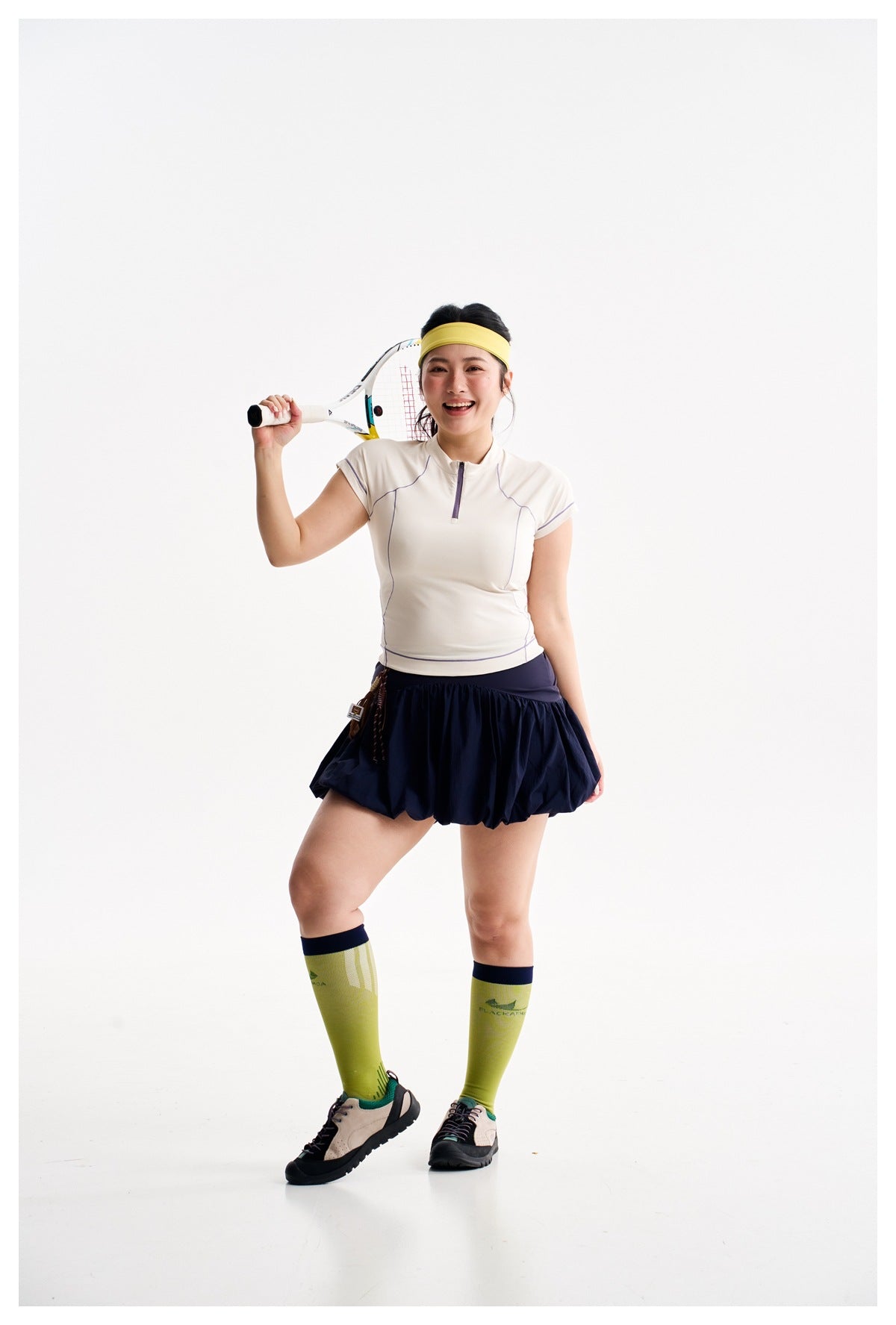 Model wearing a white cap-sleeve zip-neck top, navy bubble skirt, lime knee socks, and sneakers, holding a tennis racket.