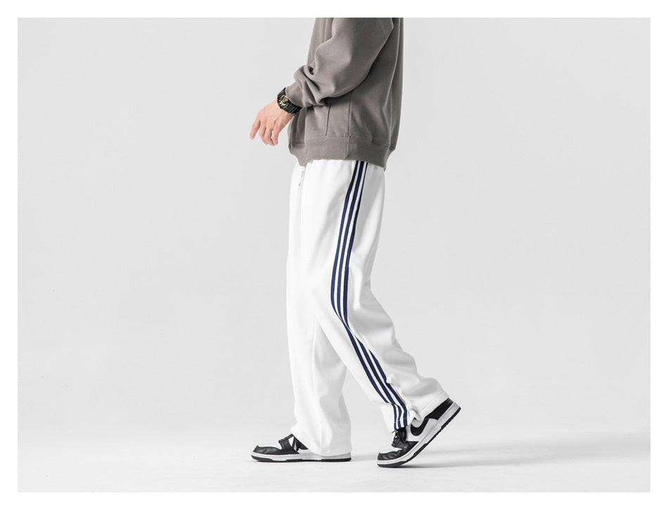 Side view of a model walking in loose white track pants with three navy side stripes and snap hems.
