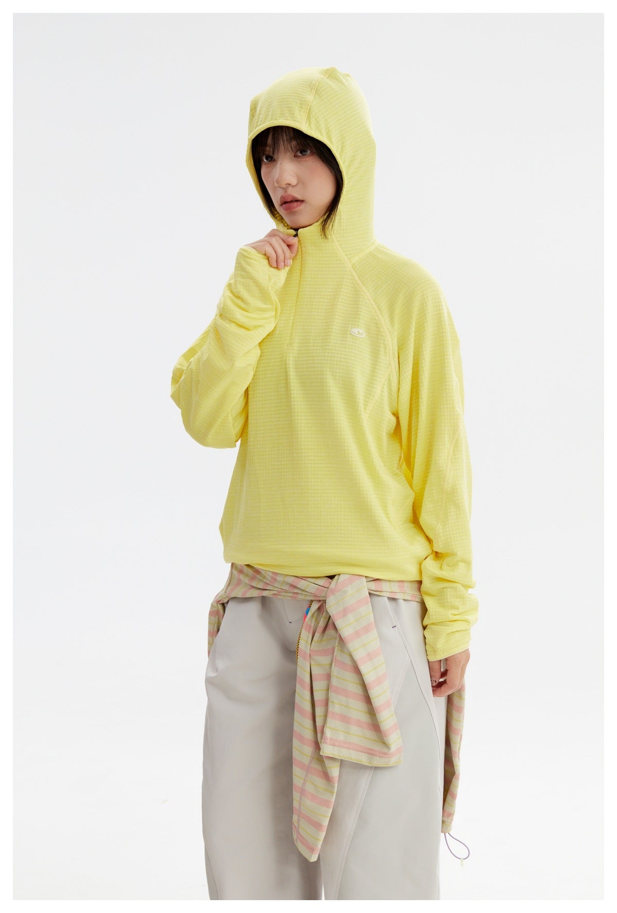 Model wearing a pale yellow hooded top with long sleeves and thumb loops, paired with light trousers.
