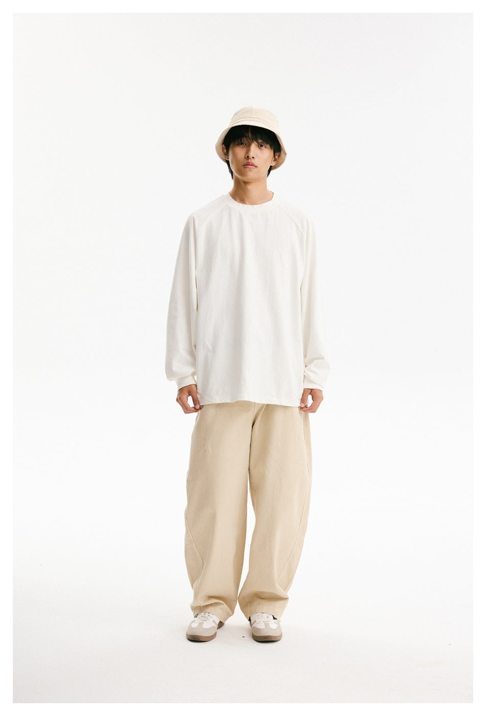 Full-length model wearing a loose white long-sleeve crewneck shirt, beige trousers, and a beige bucket hat.