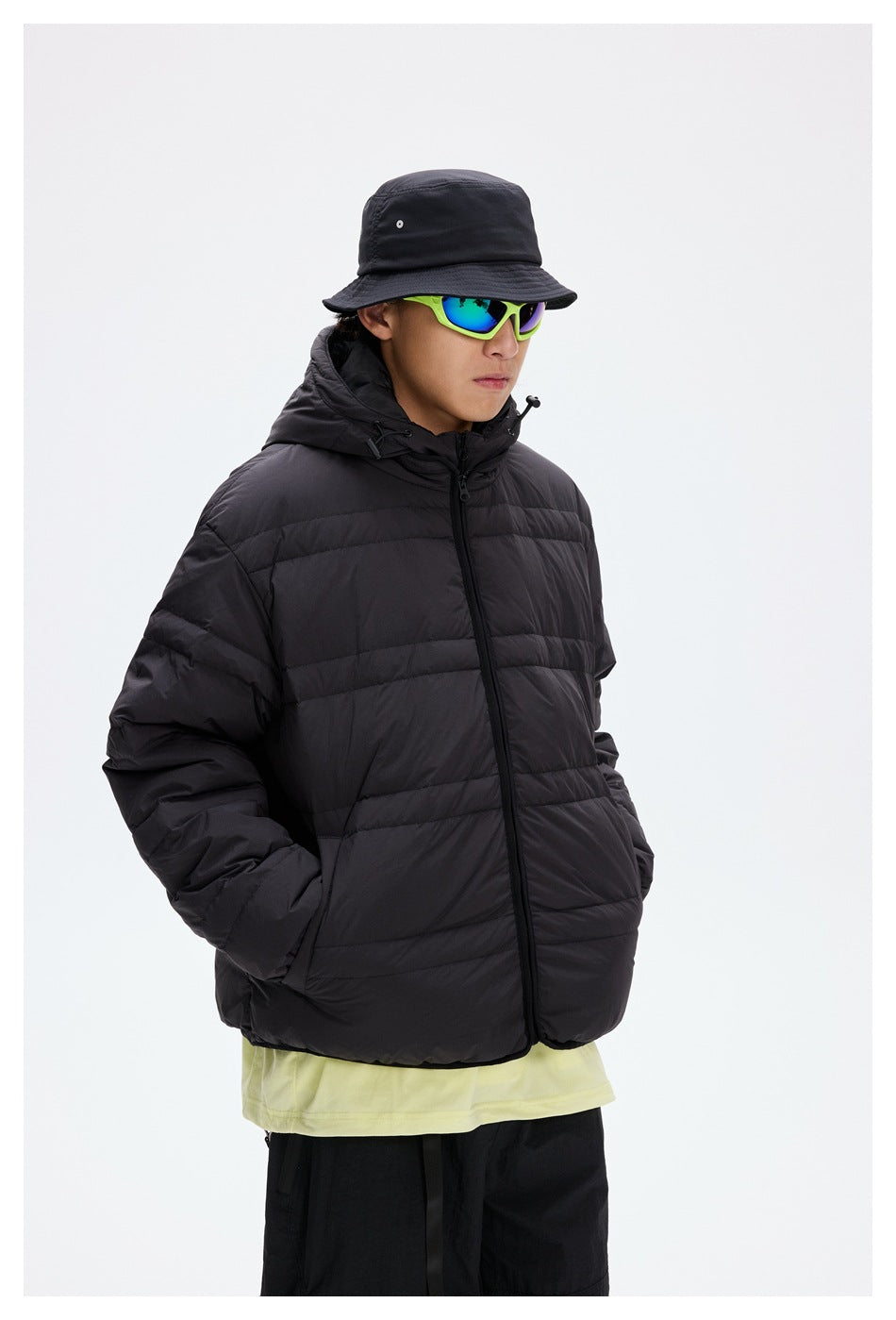Model wearing a black hooded quilted puffer jacket with a front zipper, bucket hat, and bright green sunglasses.