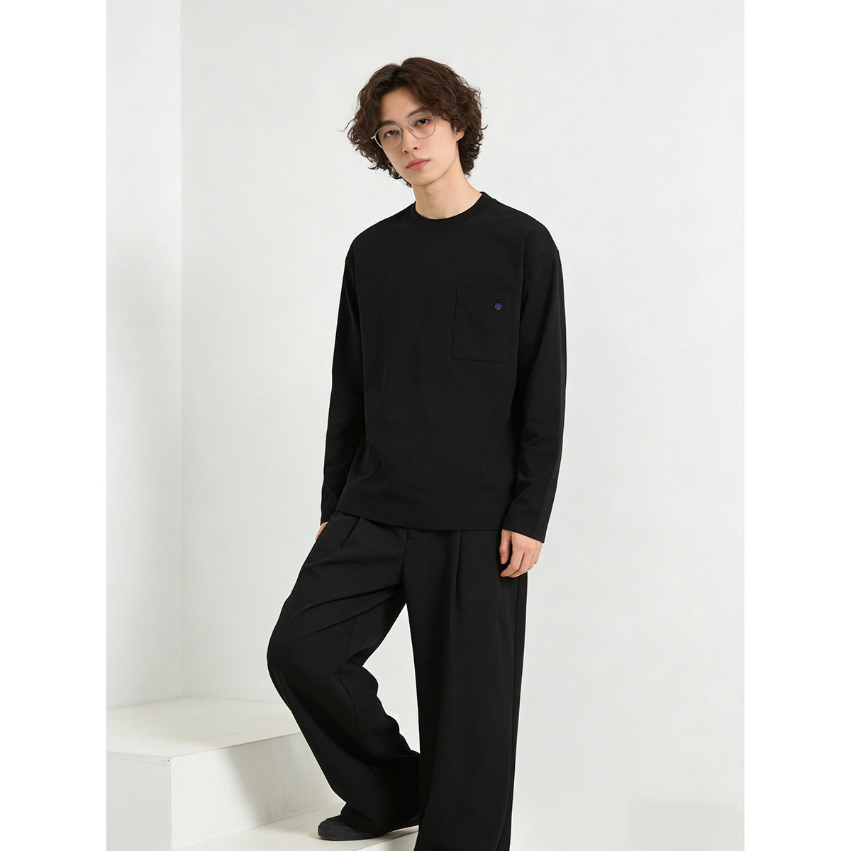 Model wearing a black long-sleeve crewneck shirt with a chest pocket and loose black trousers.