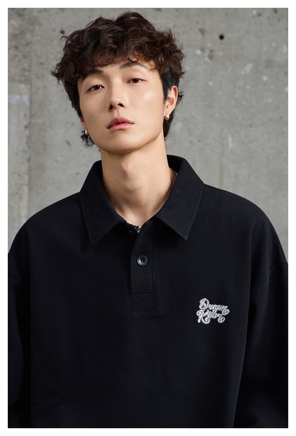Close view of a model wearing a black textured collared pullover with a button placket and white embroidered chest lettering.