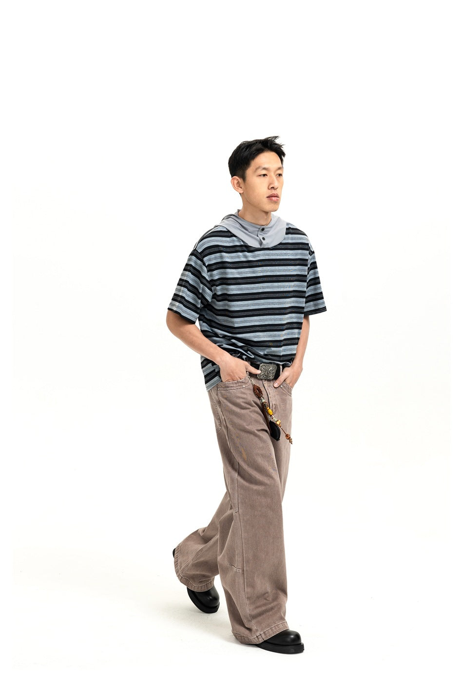 A man models a black and gray striped short-sleeve hooded shirt with loose taupe pants, in a walking pose.