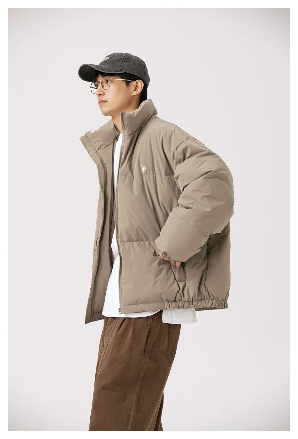 Man in a taupe-gray puffer jacket with stand collar, side view, over white tee and brown pants with a cap