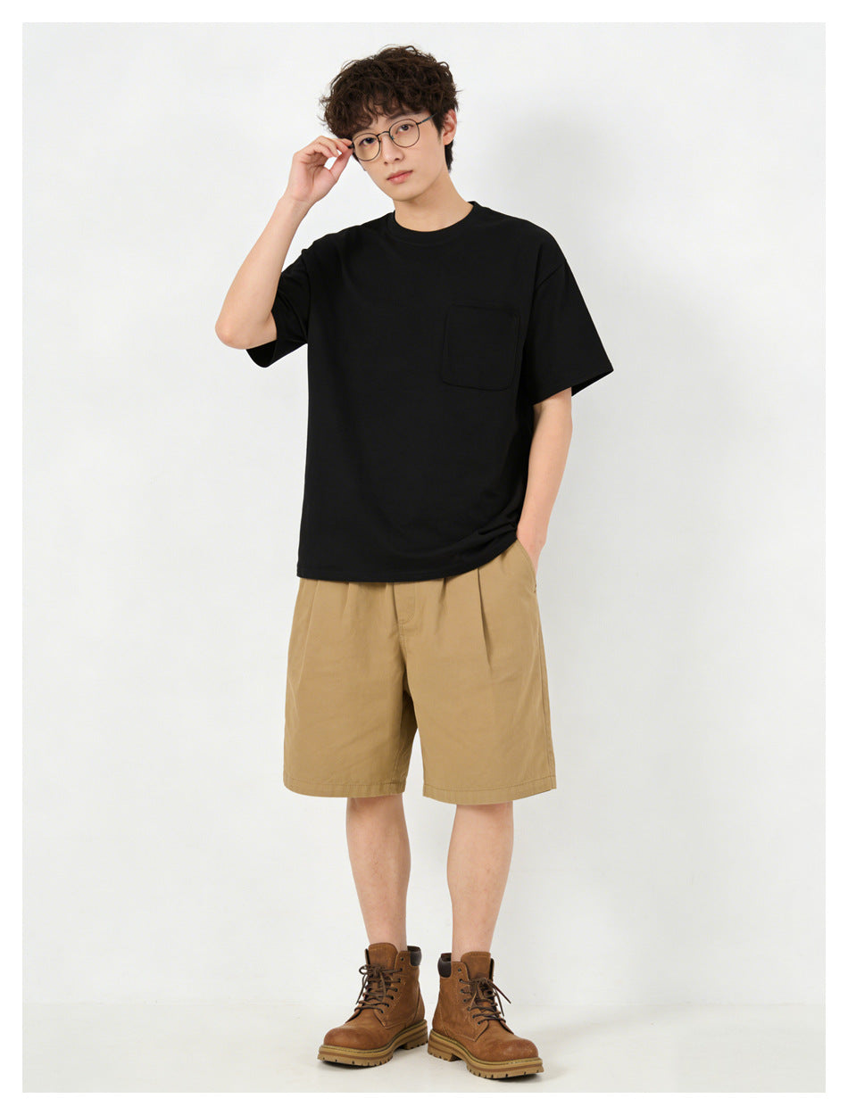 Man in black short-sleeve pocket T-shirt styled with khaki shorts and brown boots, hand at his glasses