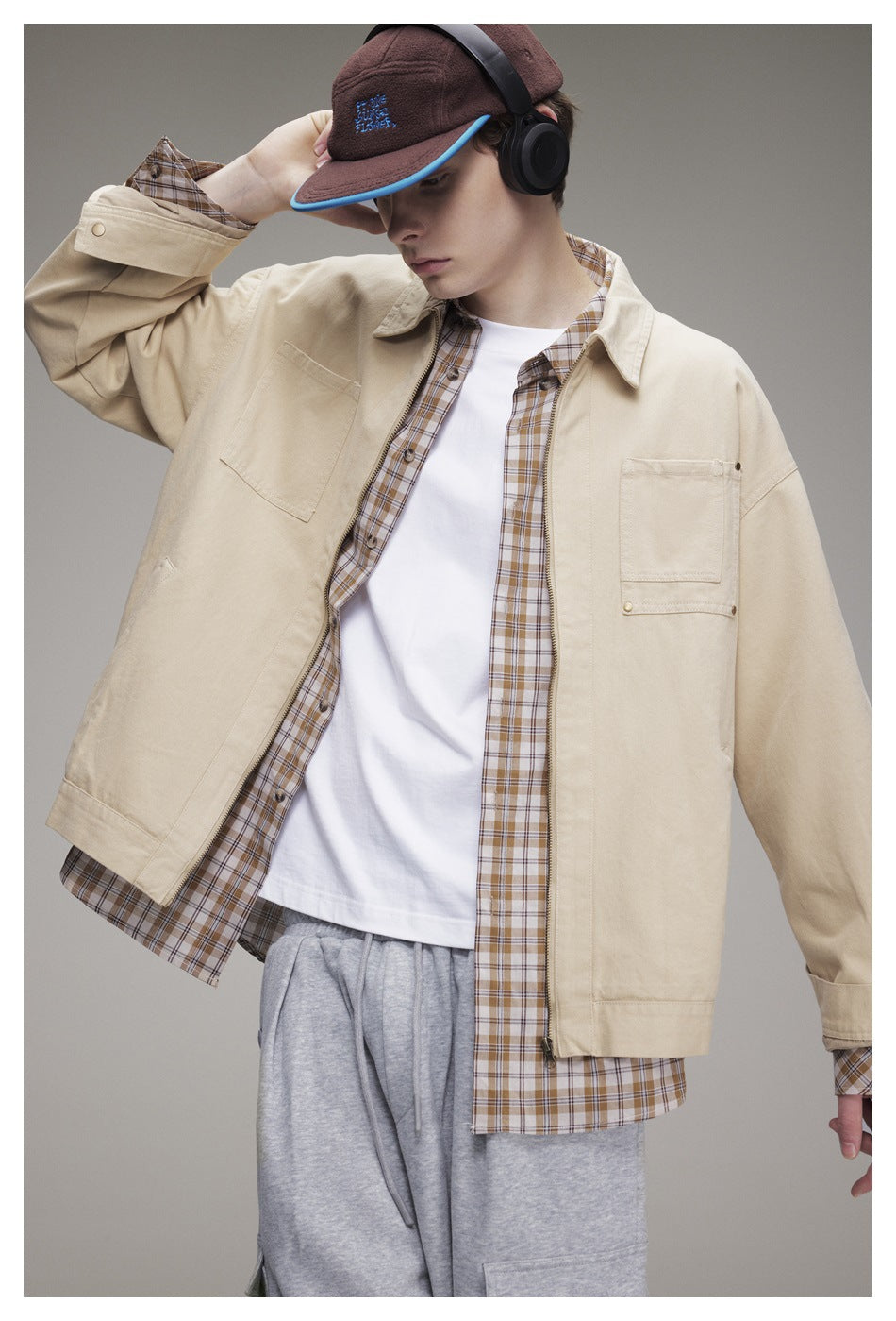 Model in a cream zip-front jacket with chest pockets, worn open over a plaid shirt and white tee.