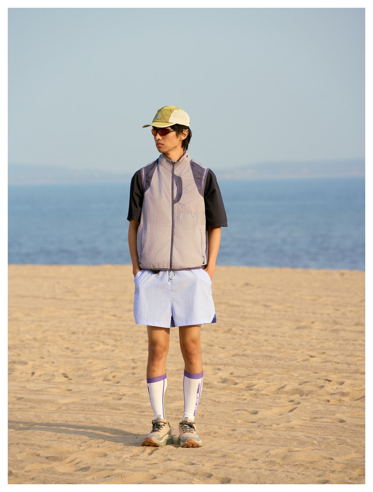 A man stands on a beach wearing a light gray utility vest over a black shirt and white shorts.