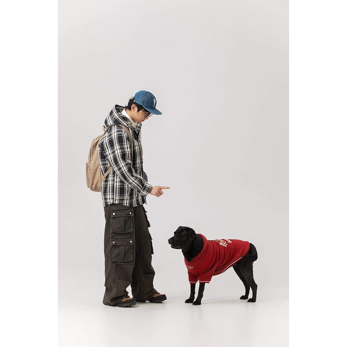 Model in a black-and-white plaid hooded jacket and dark cargo pants beside a dog wearing a red shirt.