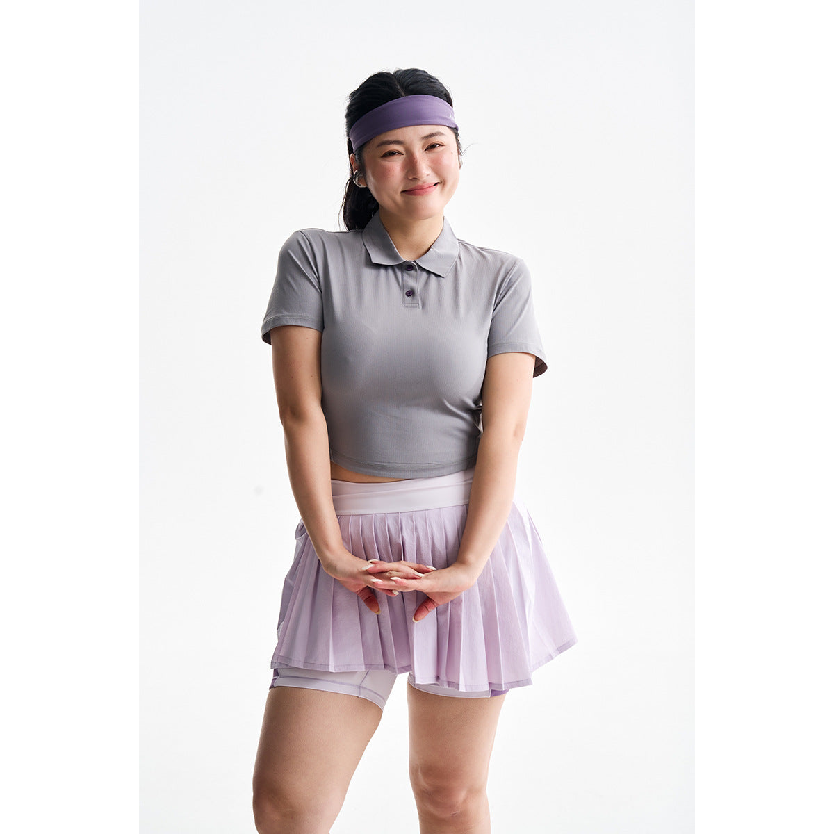 Model wearing a gray short-sleeve polo and lilac pleated tennis skirt with built-in shorts.