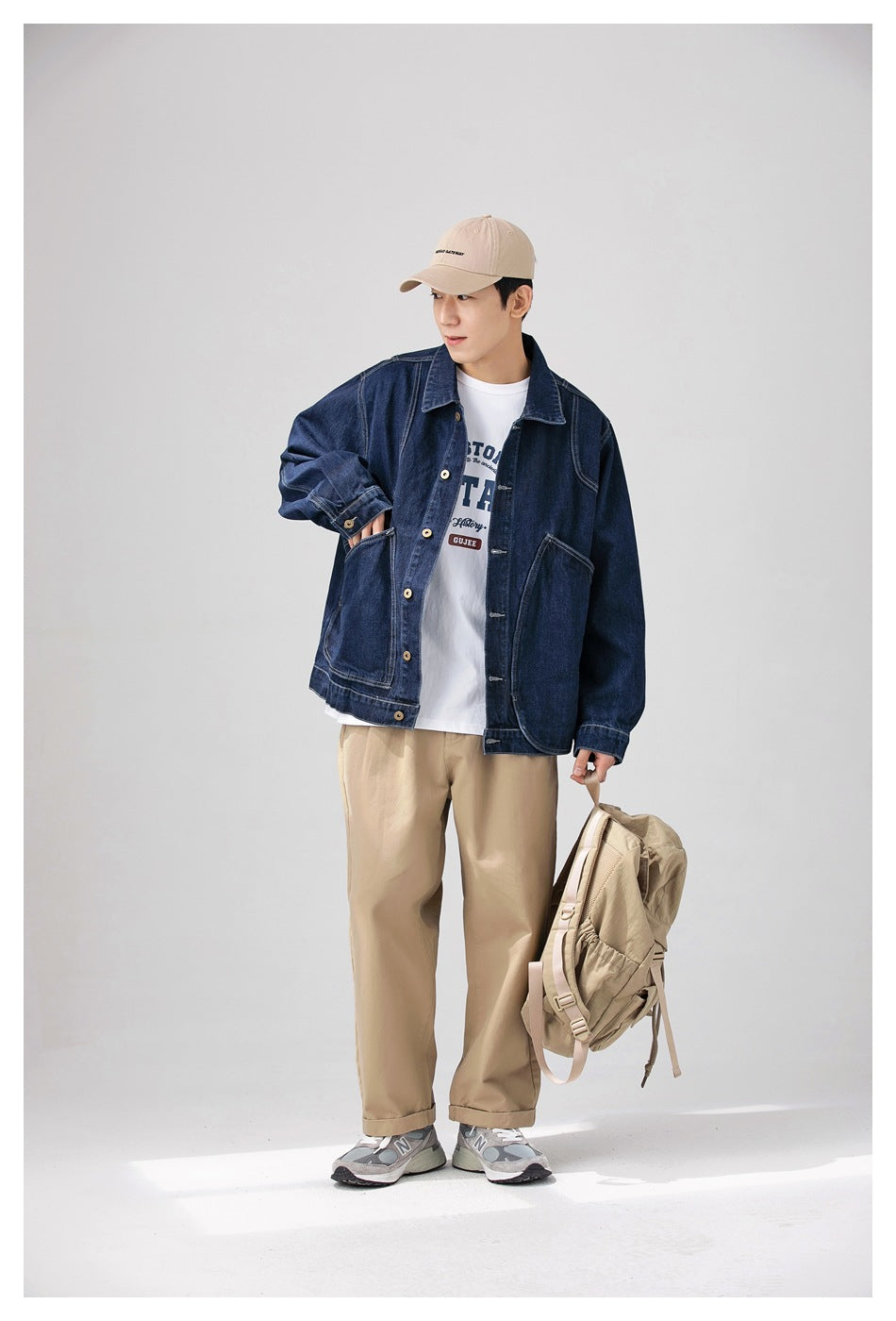 Model wearing a dark indigo denim jacket and beige trousers, with a beige backpack; white border surrounds the photo.